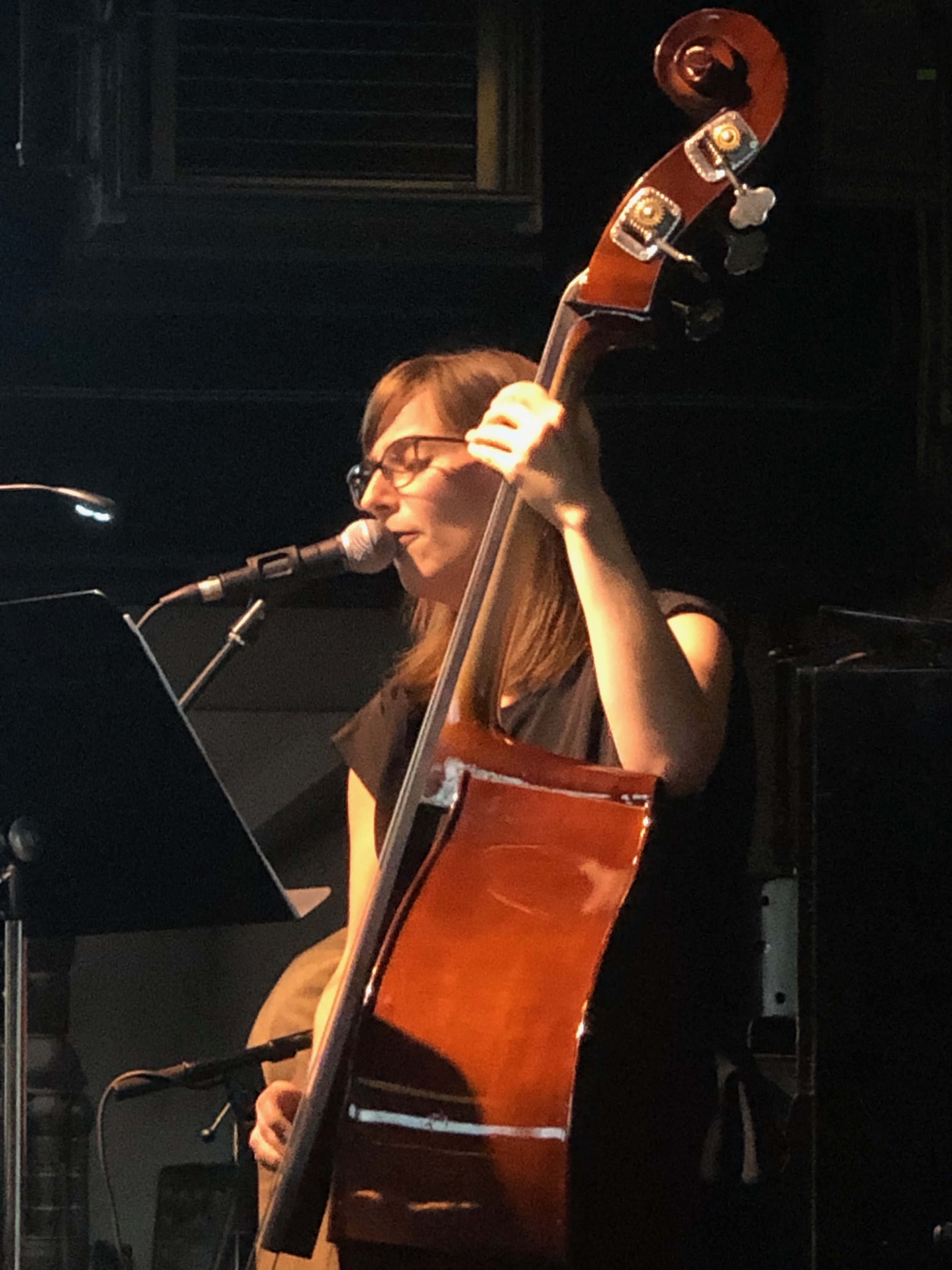 Julia sjunger i mikrofon samtidigt som hon spelar kontrabas på en mörk scen.